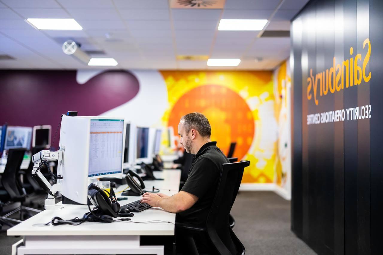 Mitie employee at a computer in the 安全 Operations Centre, with Sainsbury's branding on the walls