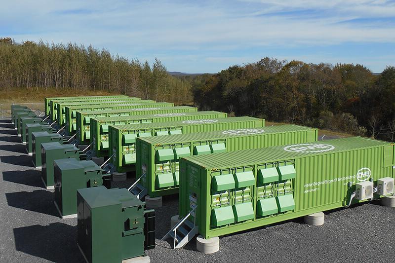 A line of large green corrugated metal containers, used for energy battery storage. They are on an asphalt surface, surrounded by trees and with a blue sky above